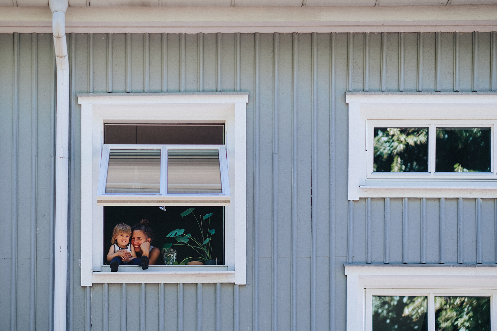 En närbild på en ljusblå husfasad med vita fönster. I ett öppet fönster kikar en mamma och hennes dotter fram.