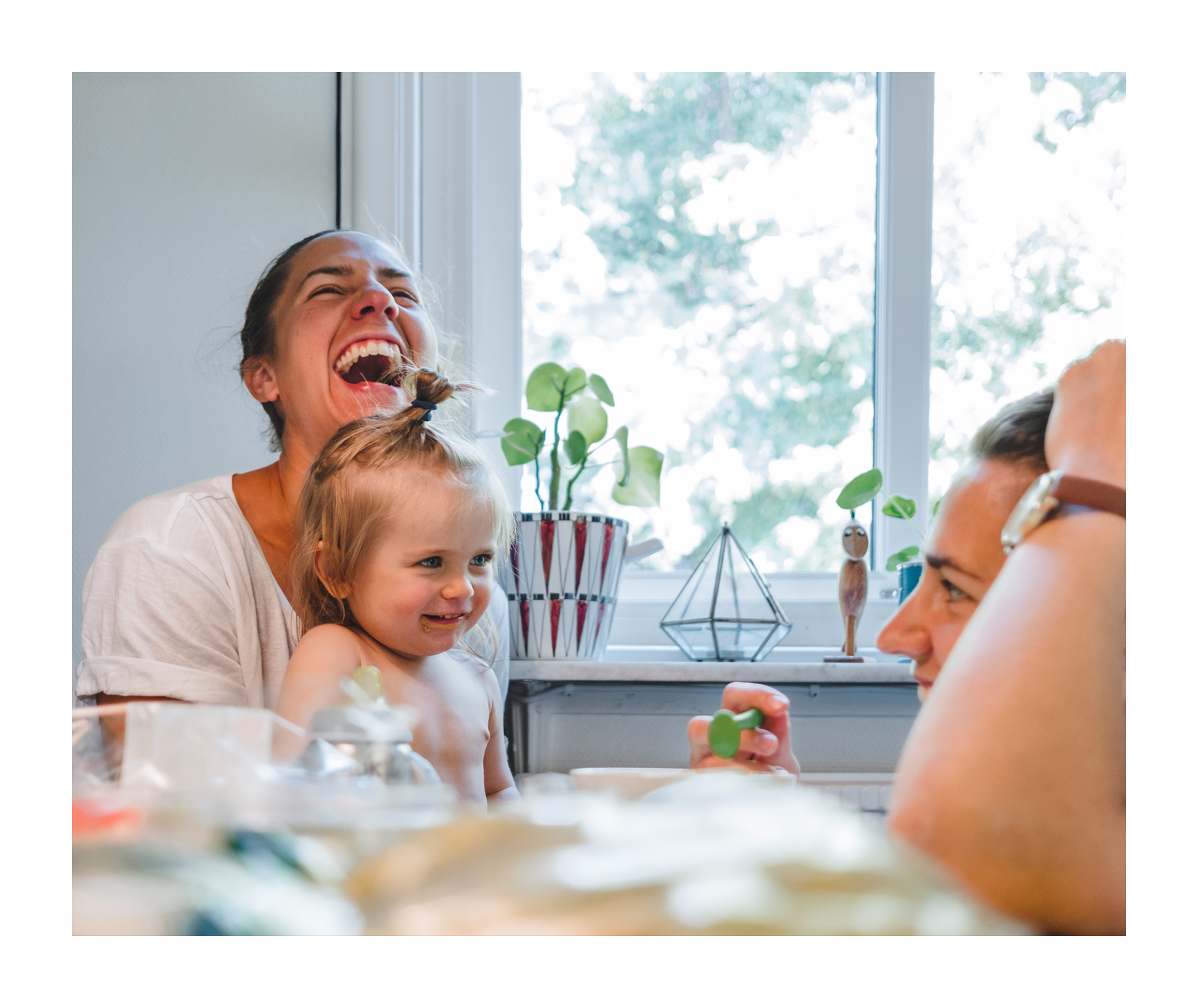 En mamma sitter med sin dotter i knät och skrattar högt. Dottern har en busig blick och tittar på en annan kvinna som sitter framför henne och ler.