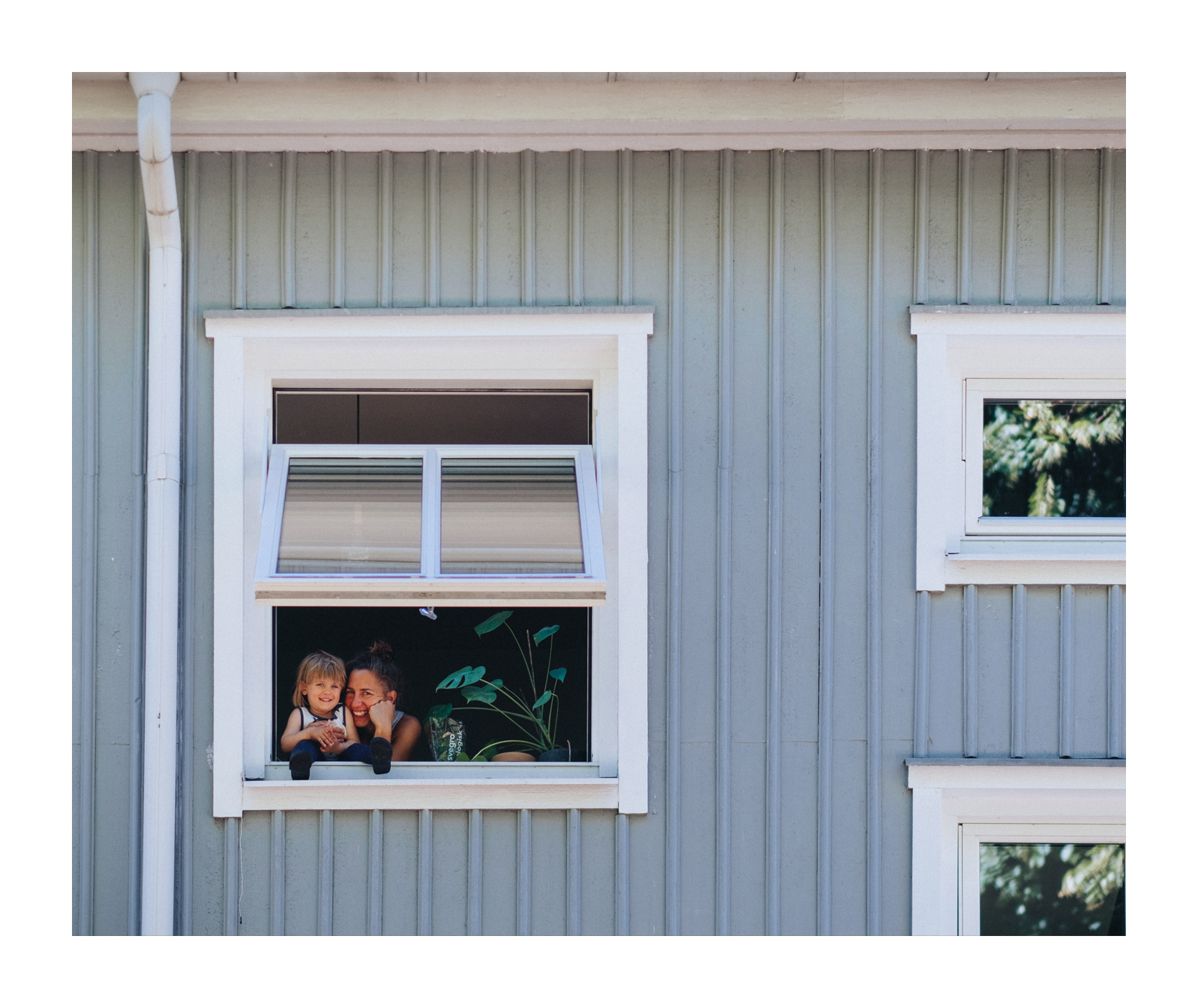 Ett trähus med ljusblå färg på fasaden och tre fönster i olika storlekar som är vita. Ett fönster är öppet och man kan se en mamma och hennes dotter skratta.