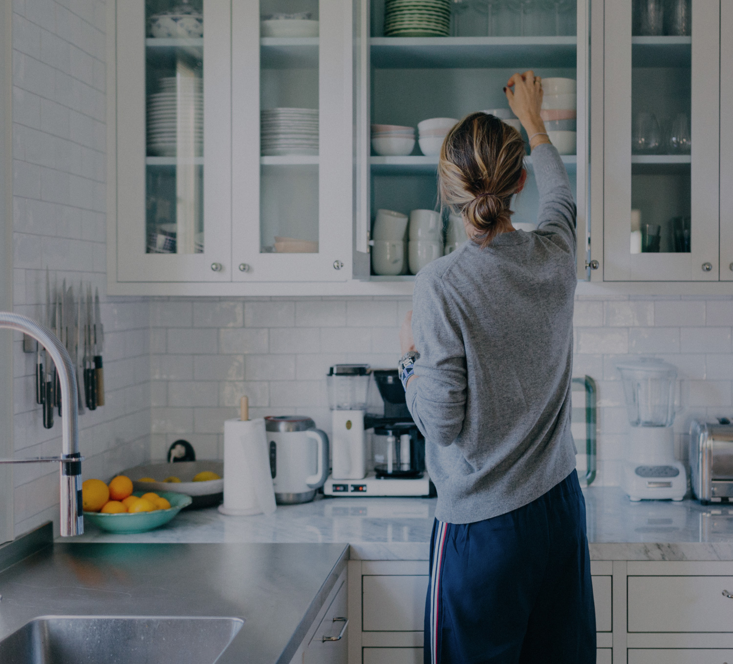 En kvinna sträcker sig mot sitt köksskåp för att ta ner en liten, djup skål. Hon har grå tröja, blå byxor med röd-vit-blå rand på sidan och håret uppsatt i en låg knut.