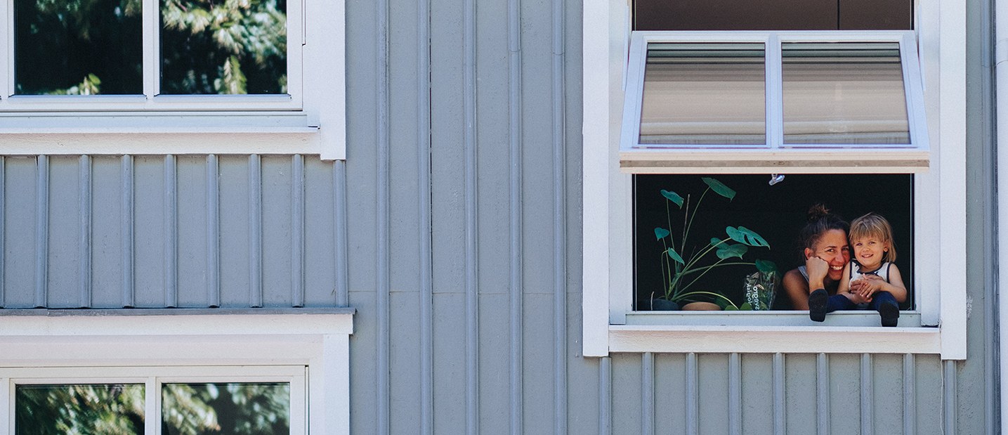 Ett trähus med ljusblå färg på fasaden och tre fönster i olika storlekar som är vita. Ett fönster är öppet och man kan se en mamma och hennes dotter skratta.
