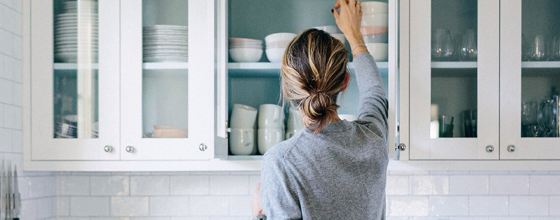 Köpa fritidshus bolån. En kvinna sträcker sig mot sitt köksskåp för att ta ner en djup skål. Hon har grå tröja och håret uppsatt i en låg knut. Köket är ljus med olika köksmaskiner ståendes på köksbänken.